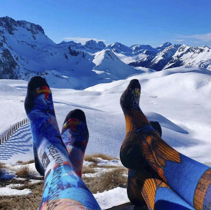 2 pairs of feet wearing smartwool ski and snowboard socks with snowy mountains in the background